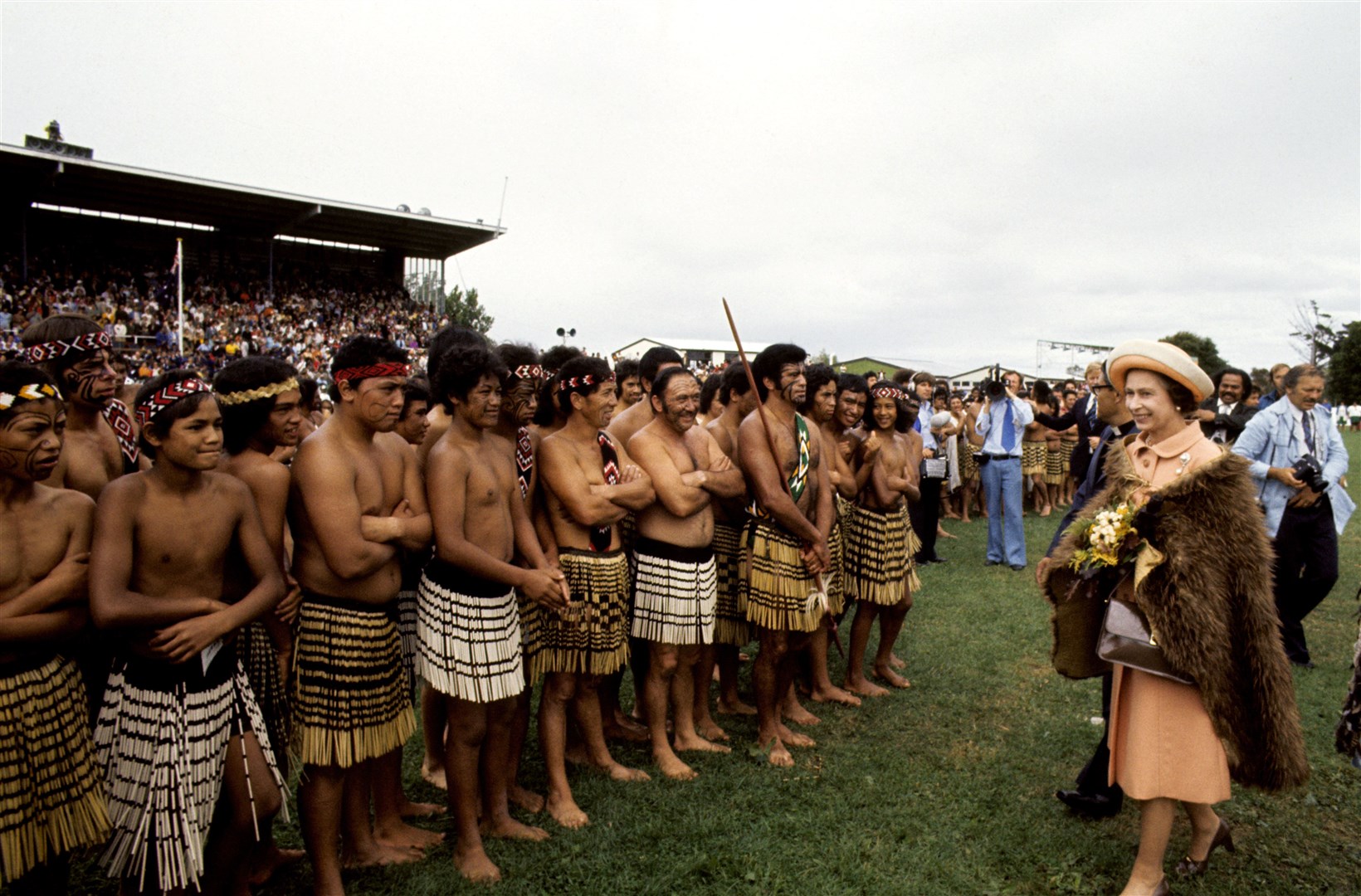 The Queen wearing a cloak of brown kiwi feathers, with Maori warriors at Rugby Park, Gisborne, New Zealand during her Silver Jubilee tour (PA)